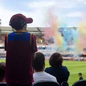 A young child in care, wearing a maroon and blue jersey, stands with hope and excitement in a stadium as colourful smoke fills the air—surrounded by supportive adults, symbolising inclusion, belonging, and brighter futures through the Little Stars Learners Program.