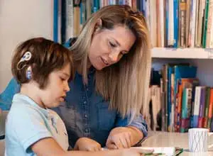 Supportive educator helping a young child with hearing aids during a learning activity at a desk, surrounded by books.
